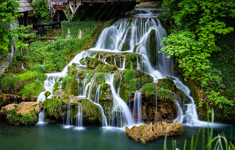 Cascading waterfall in Plitvice Lakes National Park, Croatia. Crystal-clear turquoise water flows over multiple limestone terraces surrounded by lush green forest. Wooden walkways allow visitors to explore the scenic landscape up close.