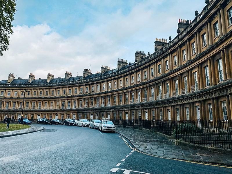 Street view of the Royal Crescent in Bath, England, showcasing its iconic sweeping arc of 30 Georgian townhouses. The uniform honey-colored stone façades face a expansive green lawn, epitomizing 18th-century urban design and elegance.