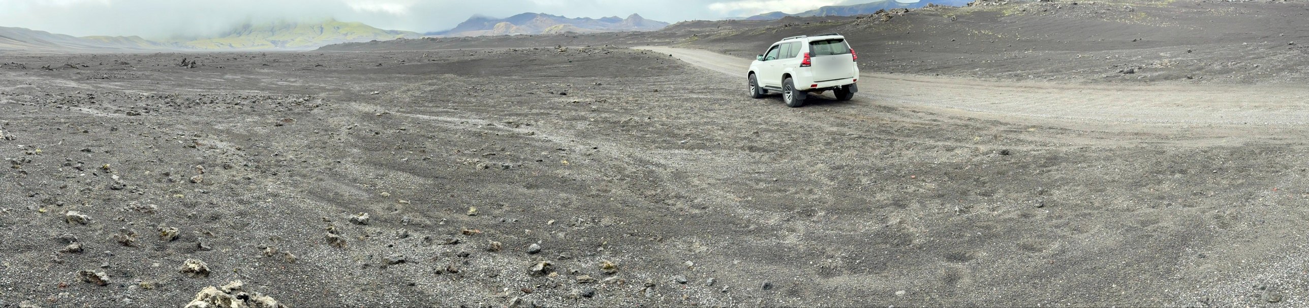 A lifted white 4x4 on a gravel F-road through grey volcanic desert, with mountains and mist beyond