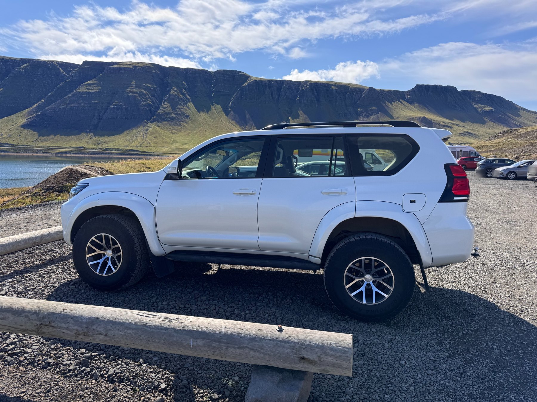 A lifted white 4x4 on large all-terrain tyres parked at a gravel car park beside a fjord